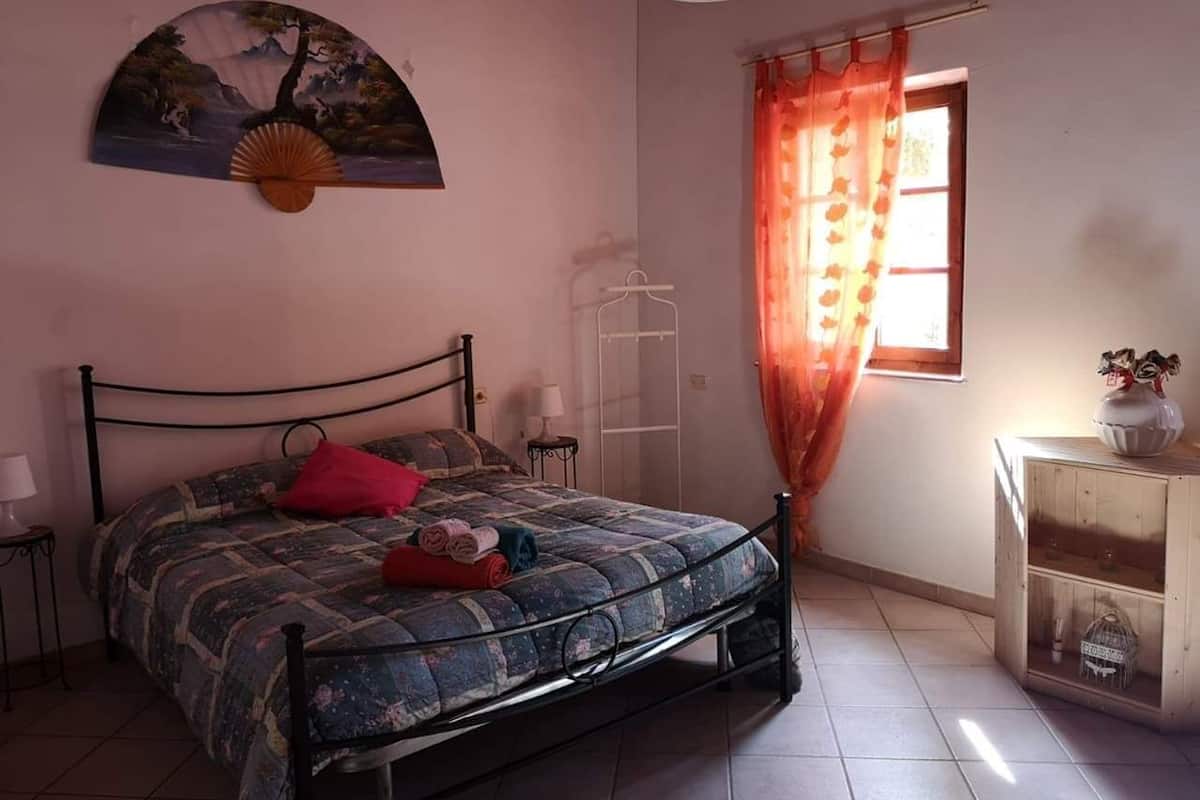 Ein Schlafzimmer mit einem Bett, einem Wandventilator, einem Fenster mit Vorhängen und einem Nachttisch mit Lampe.