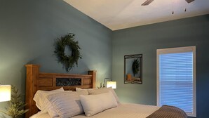Un dormitorio con cama, mesitas de noche, espejo en la pared, ventana con persianas y ventilador de techo.