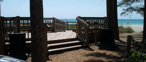 A wooden deck leading to a beach, surrounded by trees and a clear view of the ocean.