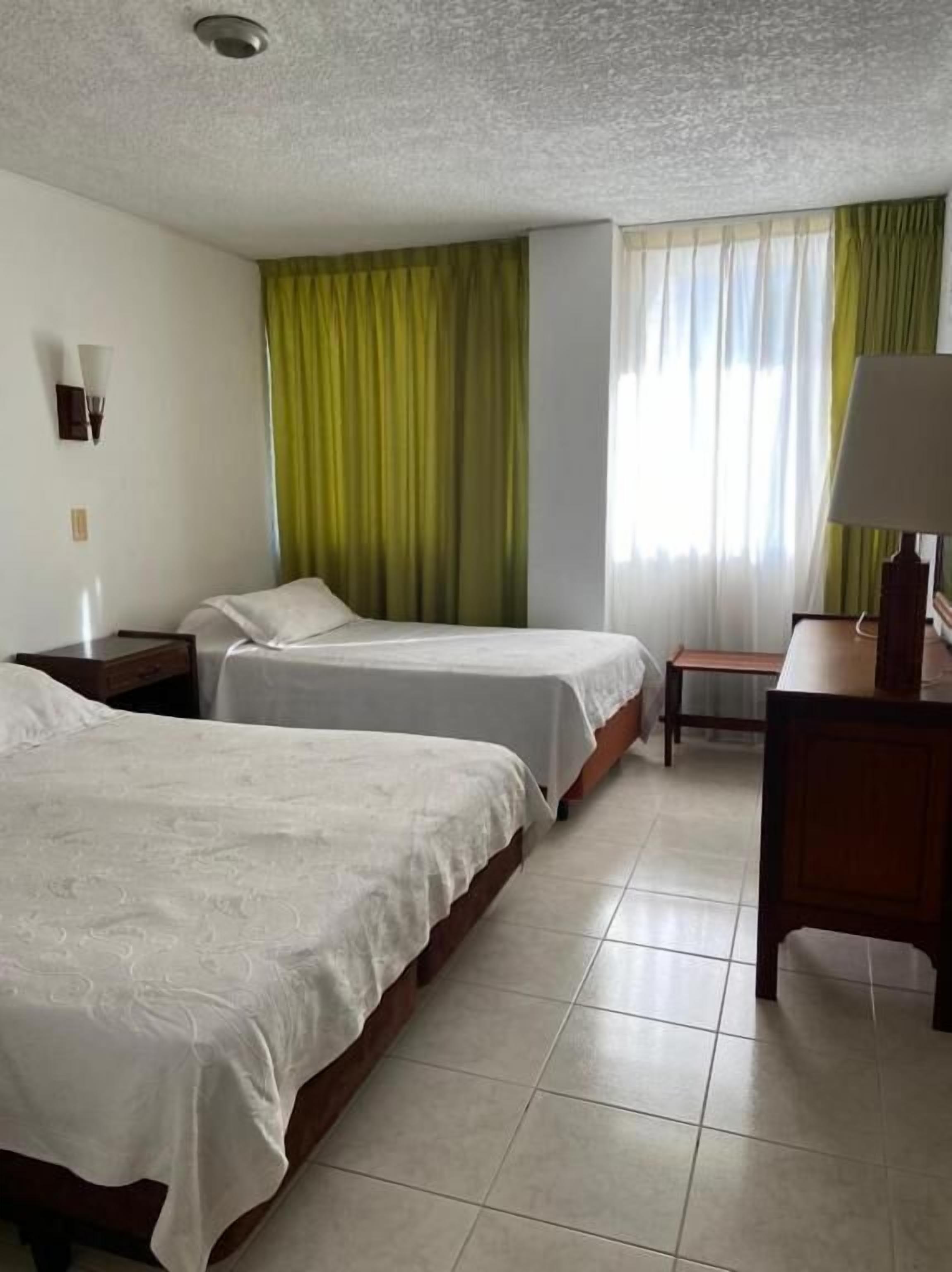 Quarto de hotel com duas camas, uma escrivaninha, uma luminária, uma mesa de cabeceira e uma janela com cortinas.