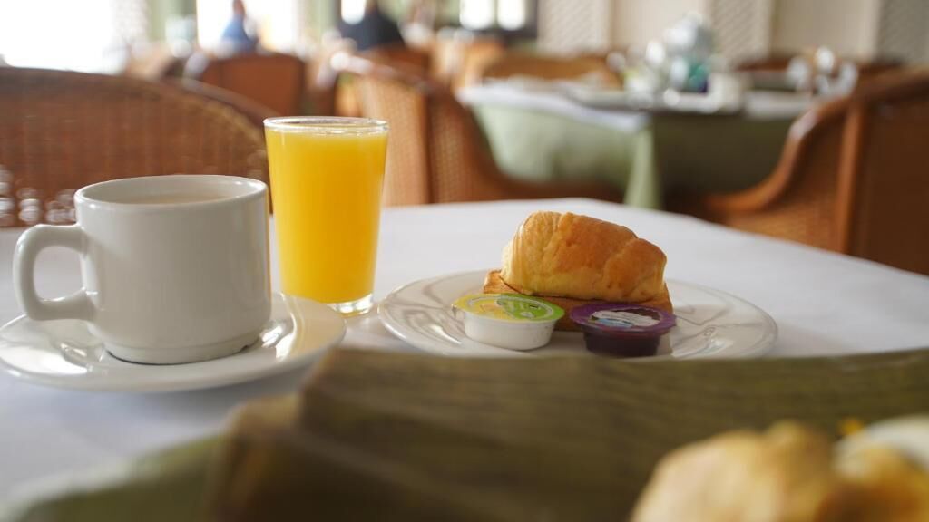 Desayuno con una taza de café, jugo de naranja, un croissant, y pequeños recipientes de mantequilla y mermelada sobre una mesa.