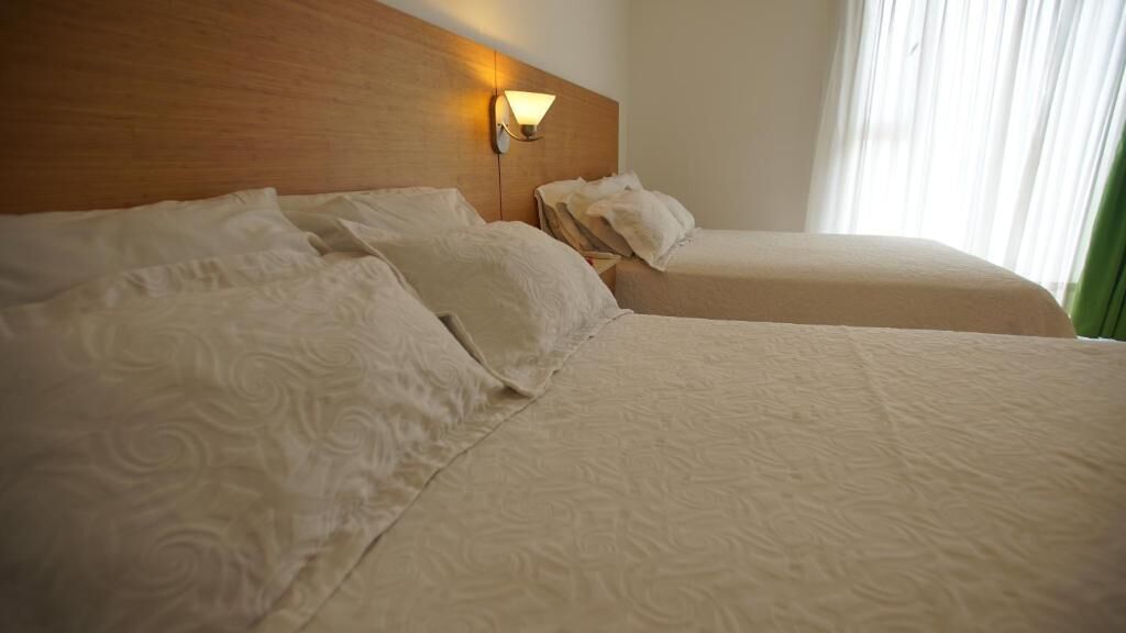 A hotel room with two beds, a wooden headboard, a wall-mounted light, and a window with curtains.