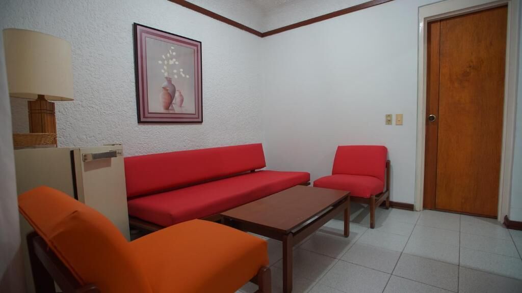 Una stanza con un divano rosso, una poltrona arancione, un tavolo in legno, una lampada e un quadro appeso al muro.