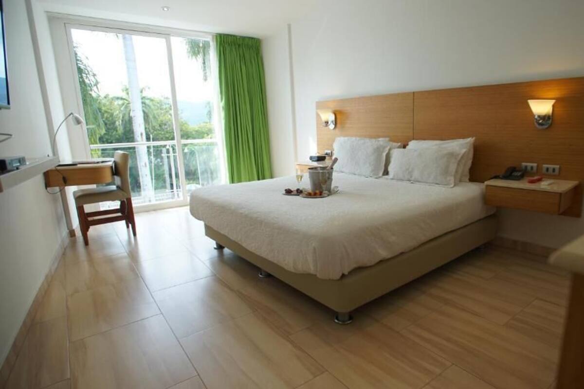 Une chambre d’hôtel avec un grand lit, un bureau, une chaise, une table de chevet et une vue sur l’extérieur grâce à une grande fenêtre.