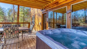 A wooden deck with a hot tub, outdoor seating, and large windows overlooking a forest.