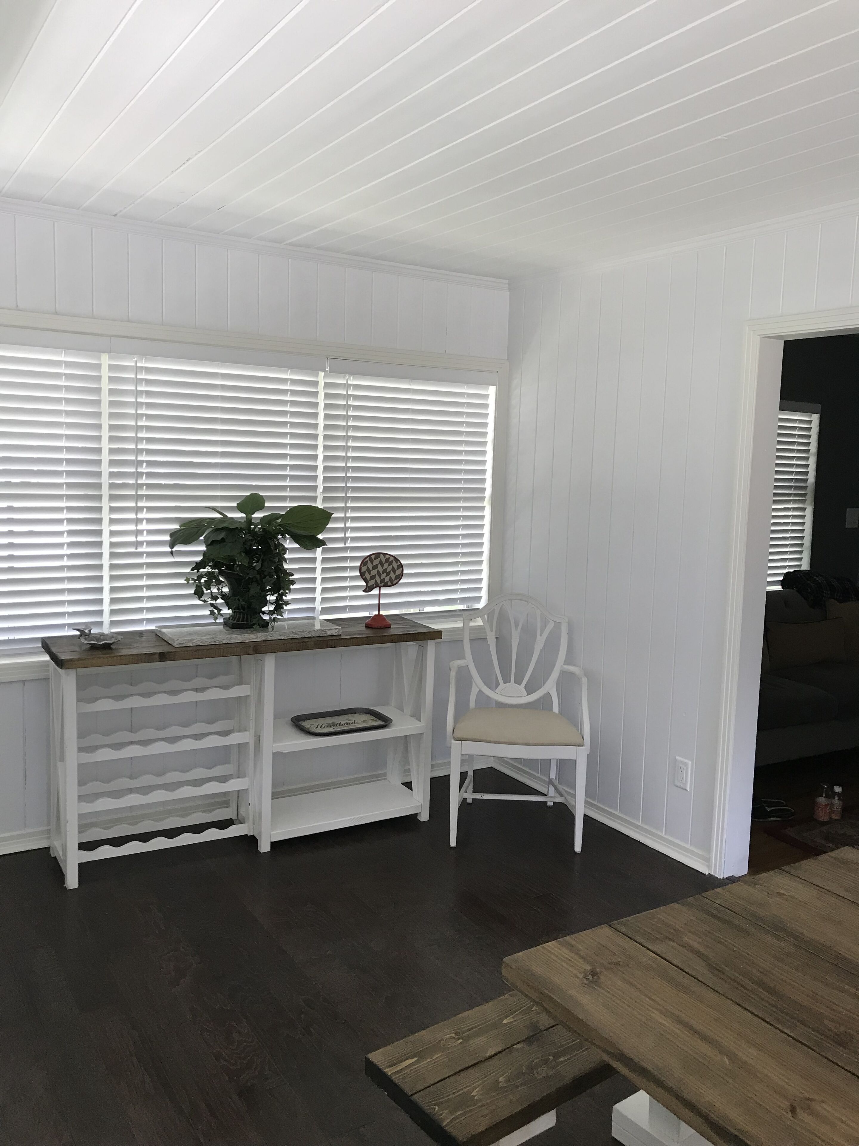 Ein Zimmer mit weißen Wandverkleidungen, einer Holzbank, einem weißen Stuhl und einem Fenster mit Jalousien.