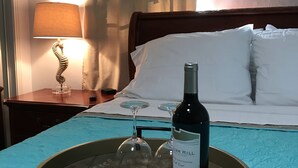 Une chambre d’hôtel avec un lit, une table de chevet avec une lampe, un plateau avec une bouteille de vin et des verres, et une fenêtre avec des rideaux.