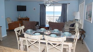 A dining area with a table set for four, surrounded by chairs. There is a television mounted on the wall and a window with curtains. - Awbrey House (Gulf Shores)