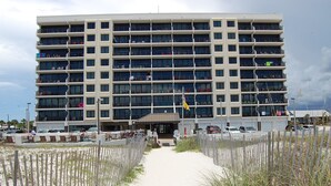 A multi-story hotel with a central entrance, surrounded by a sandy walkway and beach vegetation.