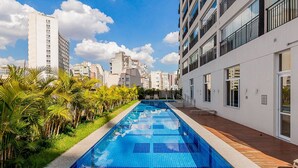 A swimming pool with a wooden deck, surrounded by palm trees and modern buildings in the background. - Autenticco - Cosmopolitan Santa Cecília (São Paulo)