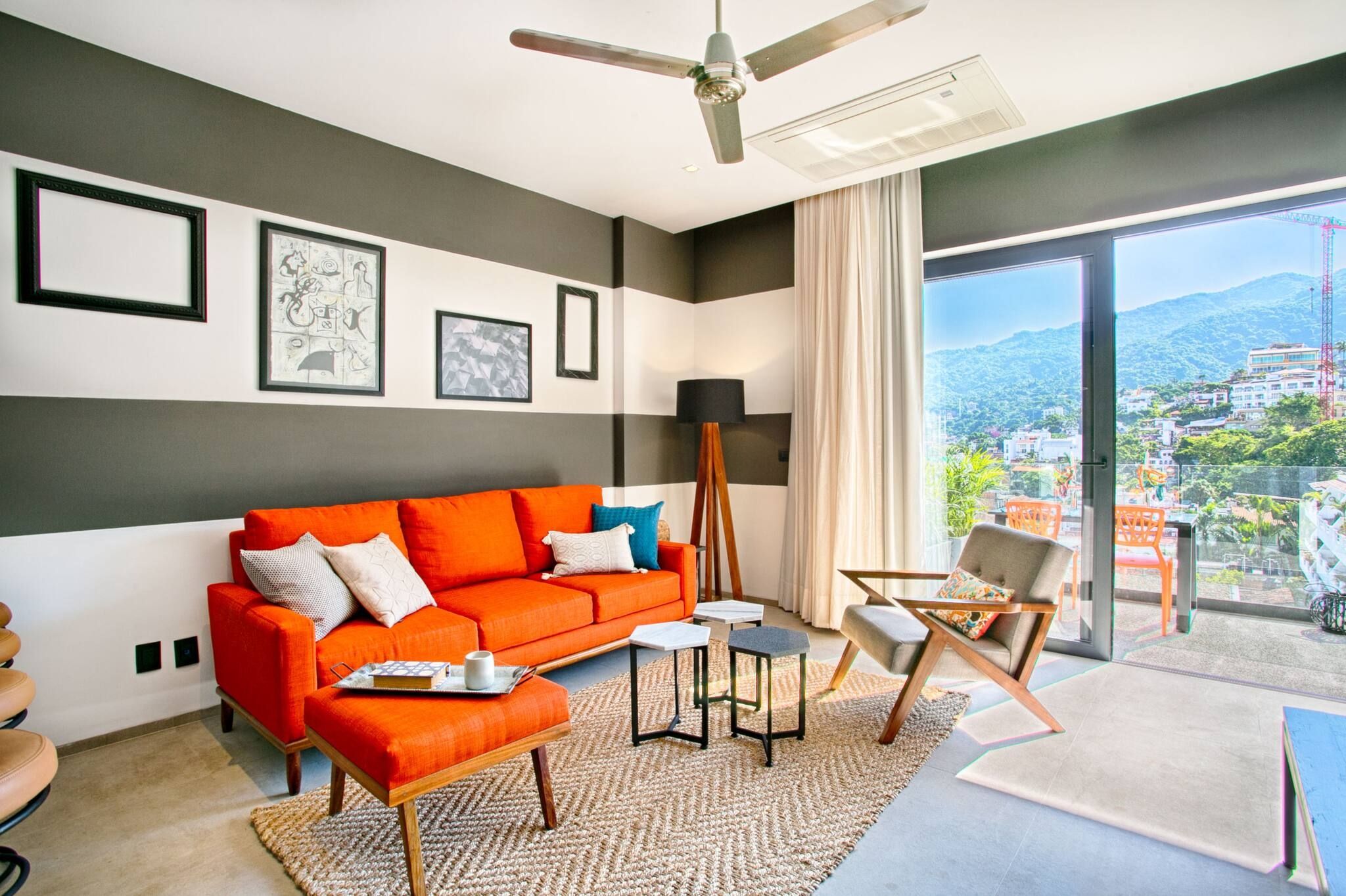 Ein modernes Wohnzimmer mit Sofa, Couchtisch und Sesseln, ein großes Fenster mit Blick auf die Stadt sowie ein Deckenventilator.