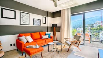 Ein modernes Wohnzimmer mit Sofa, Couchtisch und Sesseln, ein großes Fenster mit Blick auf die Stadt sowie ein Deckenventilator.