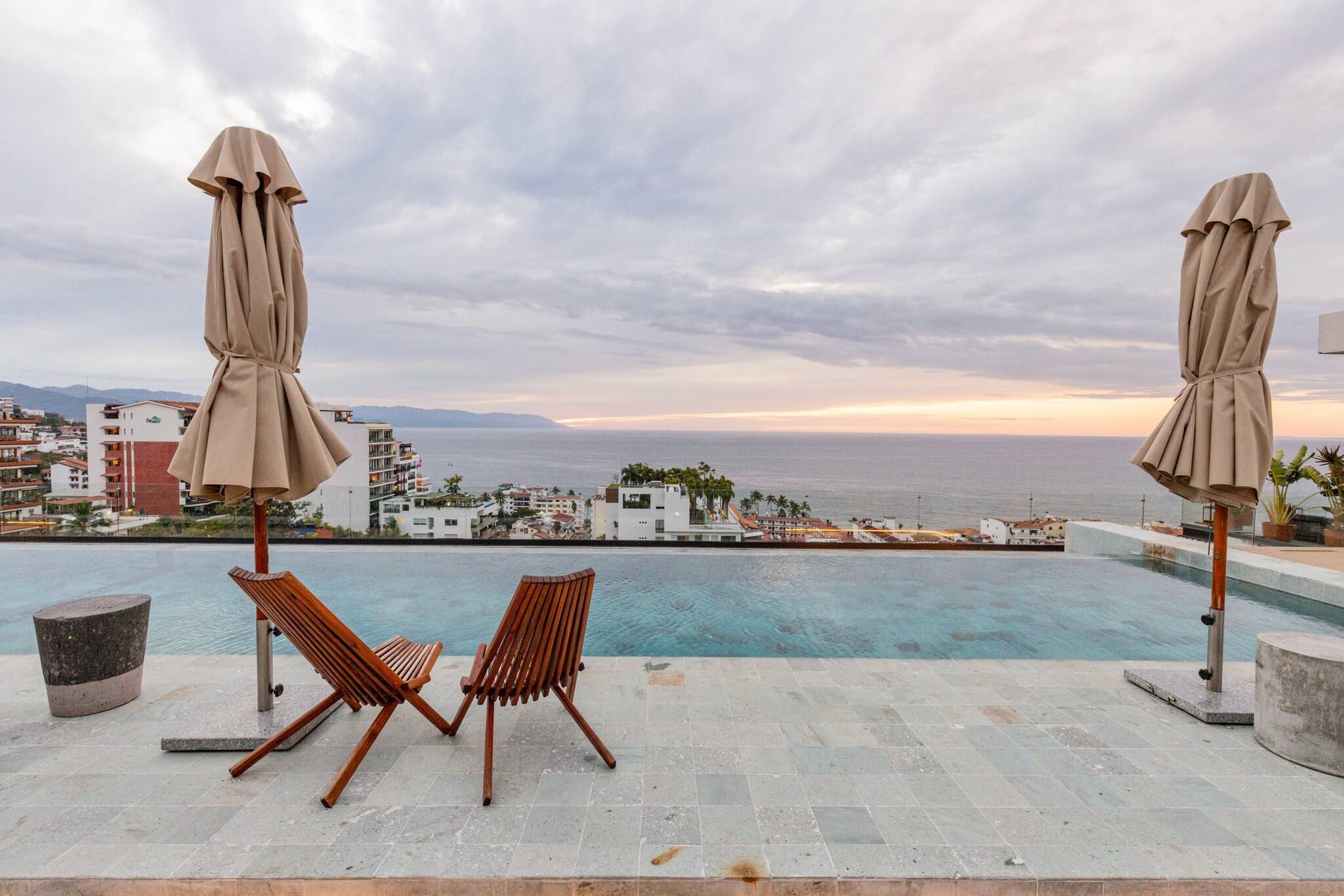 A poolside area with two wooden lounge chairs and two beige umbrellas, overlooking a coastal cityscape with buildings and a view of the sea.