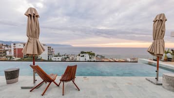 A poolside area with two wooden lounge chairs and two beige umbrellas, overlooking a coastal cityscape with buildings and a view of the sea.