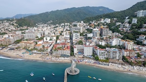 A coastal cityscape with a prominent pier extending into the sea, surrounded by densely packed buildings and a beach with numerous umbrellas. - HGTV Loft | Rooftop Pool + Gym + Views + Bar (Puerto Vallarta)