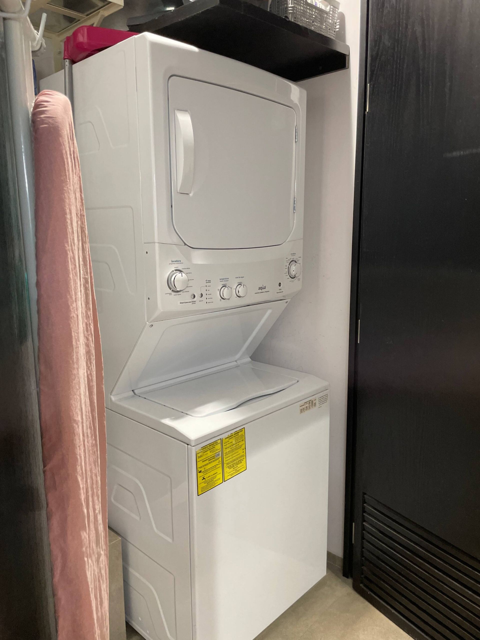 A white laundry machine with control knobs and a yellow warning label, located next to a black refrigerator in a kitchen.