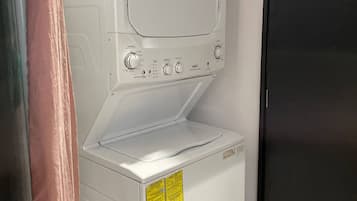 A white laundry machine with control knobs and a yellow warning label, located next to a black refrigerator in a kitchen.