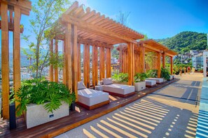 A wooden pergola with outdoor seating, surrounded by greenery and overlooking a cityscape. - HGTV Loft | Rooftop Pool + Gym + Views + Bar (Puerto Vallarta)