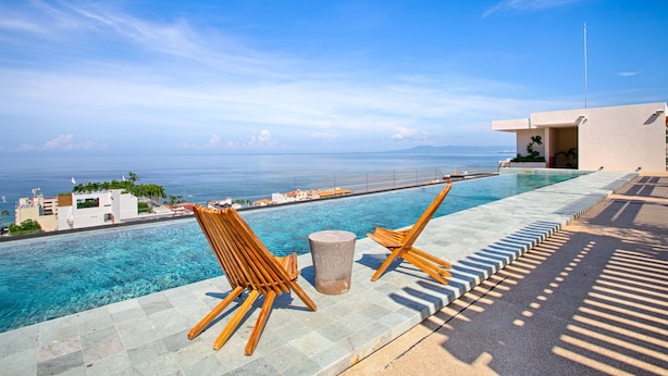 A poolside area with two wooden lounge chairs, a stone stool, and a clear view of the ocean and distant buildings. - HGTV Loft | Rooftop Pool + Gym + Views + Bar (Puerto Vallarta)