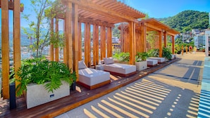A wooden pergola with seating and planters, overlooking a cityscape with buildings and hills. - HGTV Loft | Rooftop Pool + Gym + Views + Bar (Puerto Vallarta)