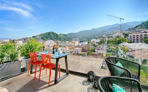 A terrace with a table and chairs, overlooking a cityscape with buildings and mountains. - HGTV Loft | Rooftop Pool + Gym + Views + Bar (Puerto Vallarta)