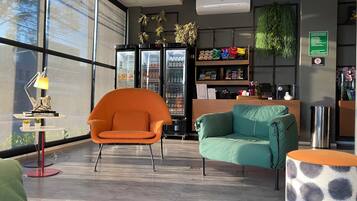 A modern lobby with a seating area featuring an orange chair and a teal sofa, a refrigerator stocked with beverages, and a small table with a lamp.