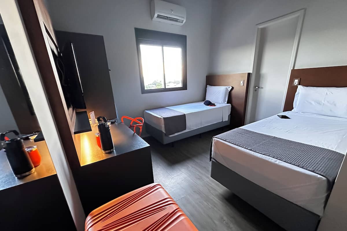 Habitación de hotel con cama, escritorio con silla, mesita con tetera, ventana y televisor.