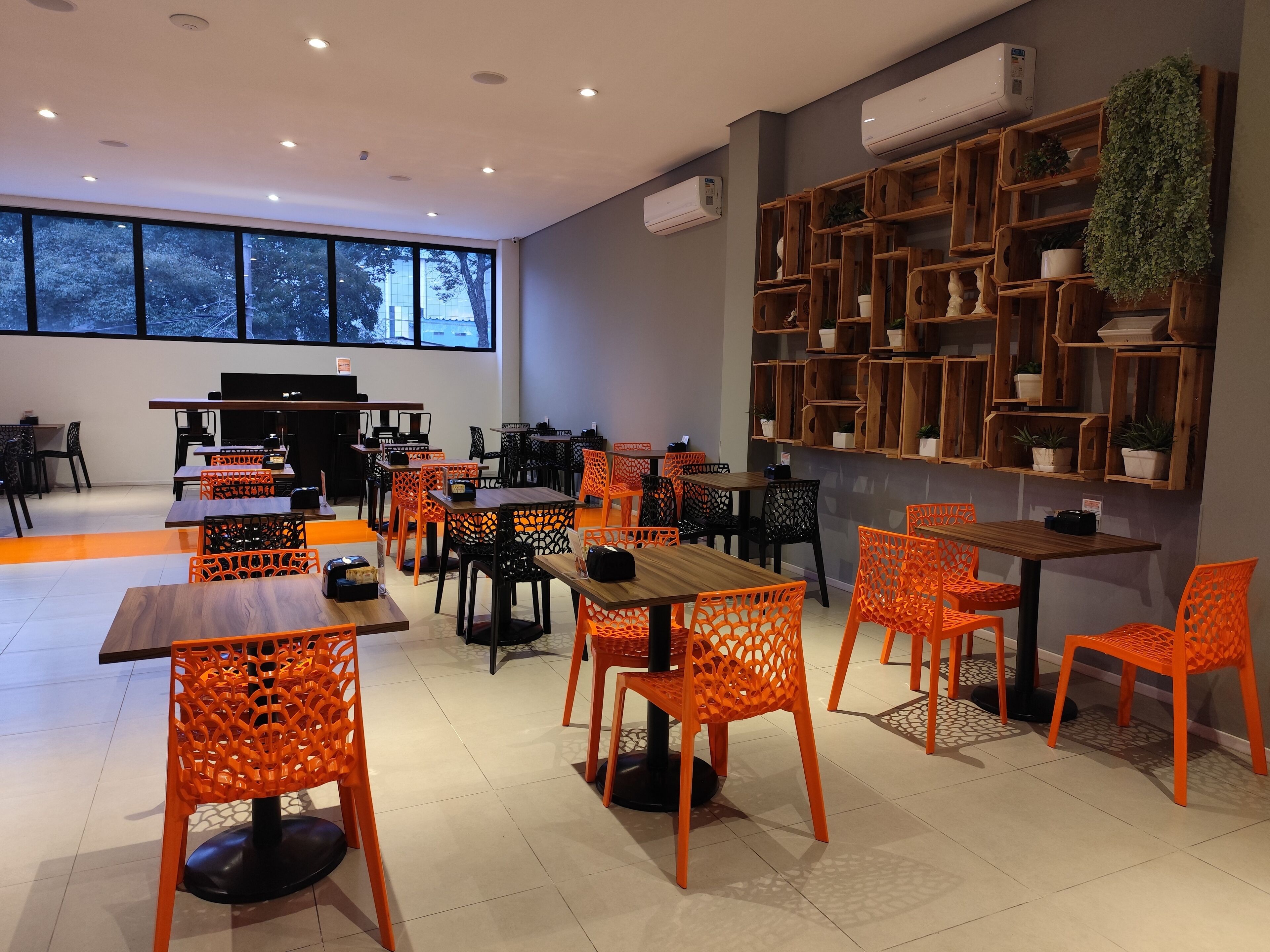 A modern indoor space with multiple tables and chairs, a large window with a view of trees, and wooden shelves with plants.