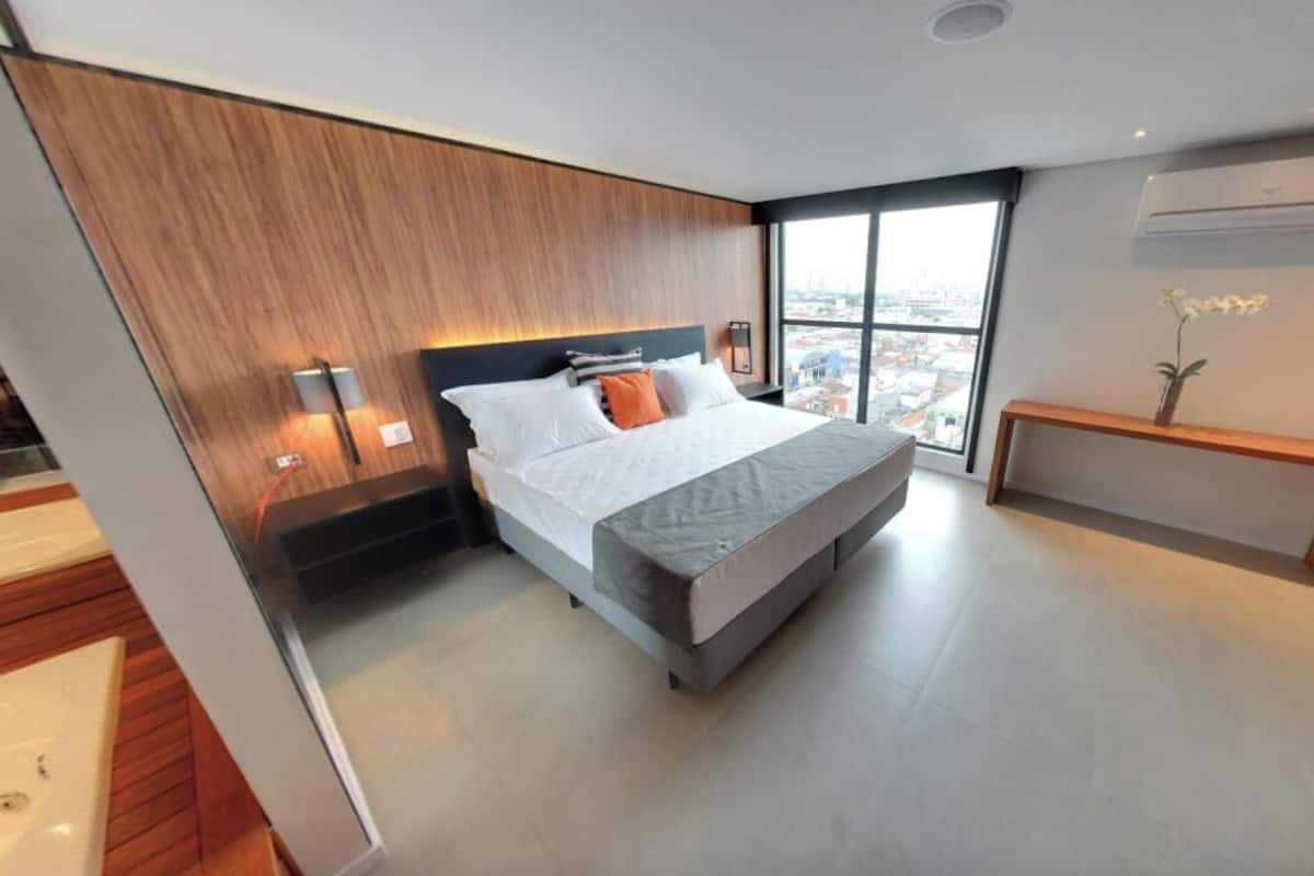 Un dormitorio moderno con una cama grande, una mesita de noche, un escritorio pequeño y una silla, y una ventana con vistas a la ciudad.
