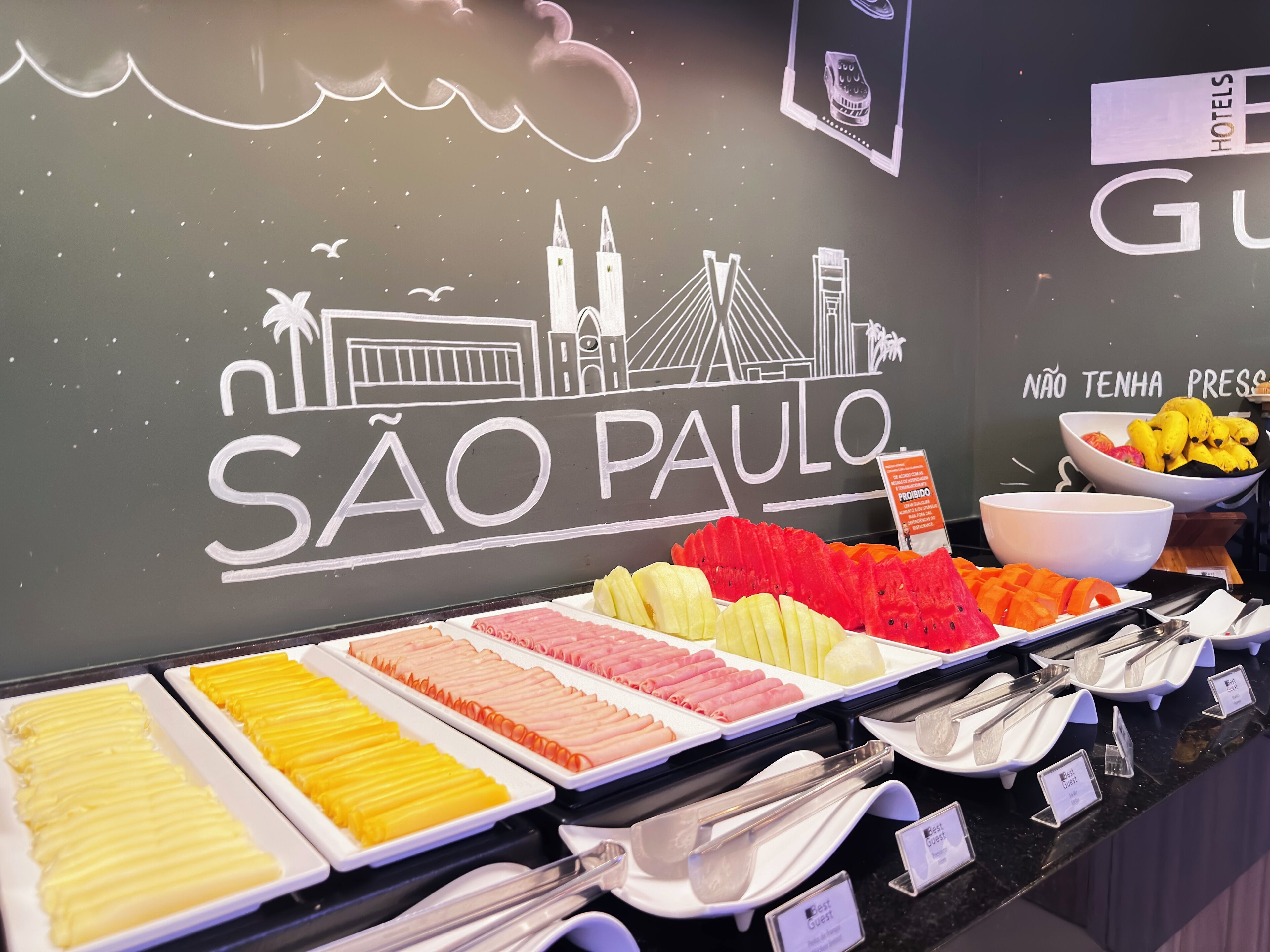 A buffet with various food items and a wall with the text "SÃO PAULO" and a cityscape illustration.