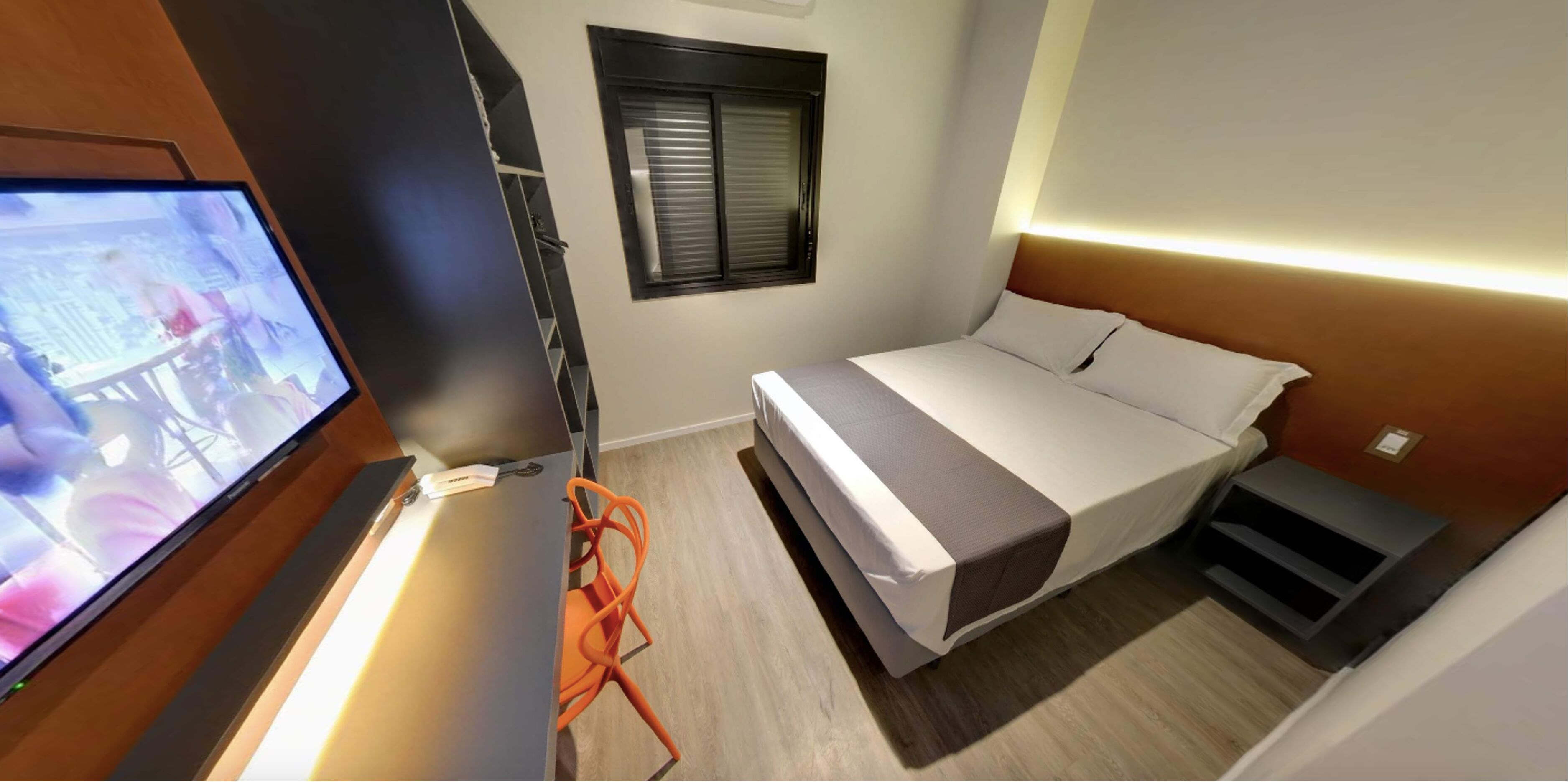 Un dormitorio con cama, televisor, silla, ventana y mesita de noche.