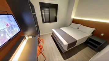 Un dormitorio con cama, televisor, silla, ventana y mesita de noche.
