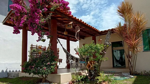 A covered patio area with a pergola, a small garden, and a house with a visible entrance. - Beautiful 3 bedroom house with private pool in a gated community close to the beach   (Jacuípe)