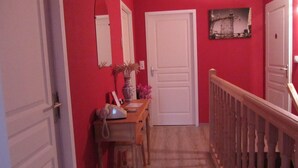 Um corredor com paredes vermelhas, uma porta branca, uma mesa de madeira com telefone, um vaso com plantas secas e um quadro na parede.