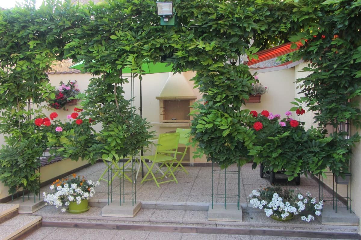 A covered outdoor seating area with a green metal chair and a small table, surrounded by lush greenery and flowering plants.