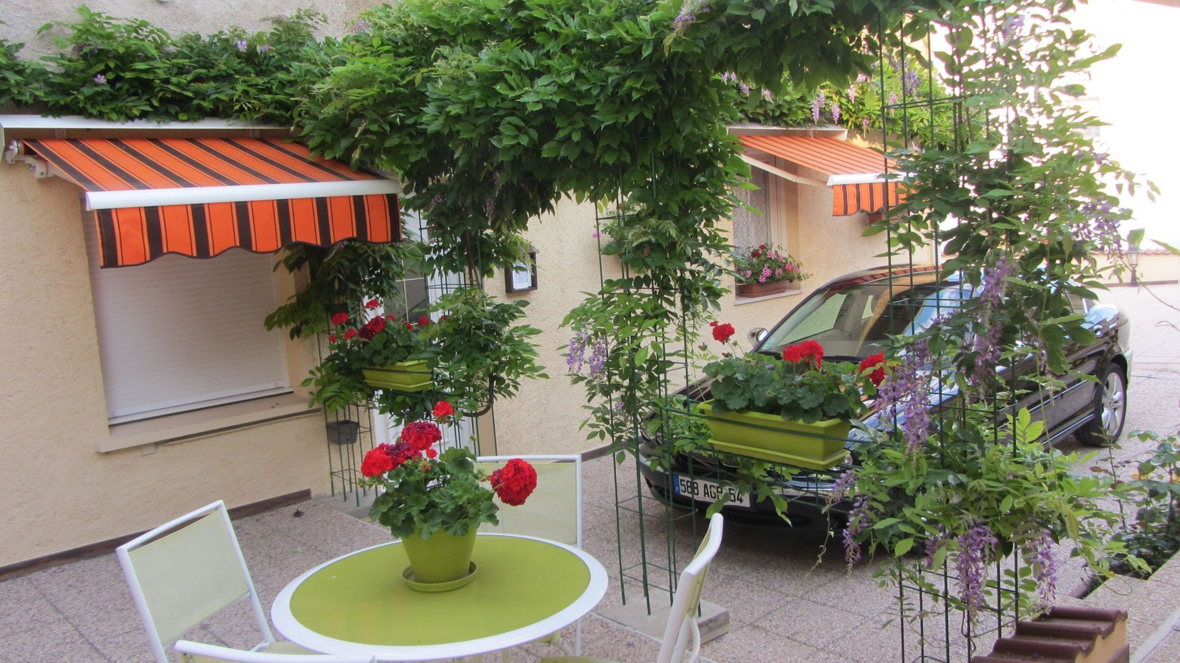 A small outdoor seating area with a table and chairs, surrounded by potted plants and a car parked nearby.