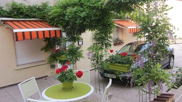 A small outdoor seating area with a table and chairs, surrounded by potted plants and a car parked nearby.