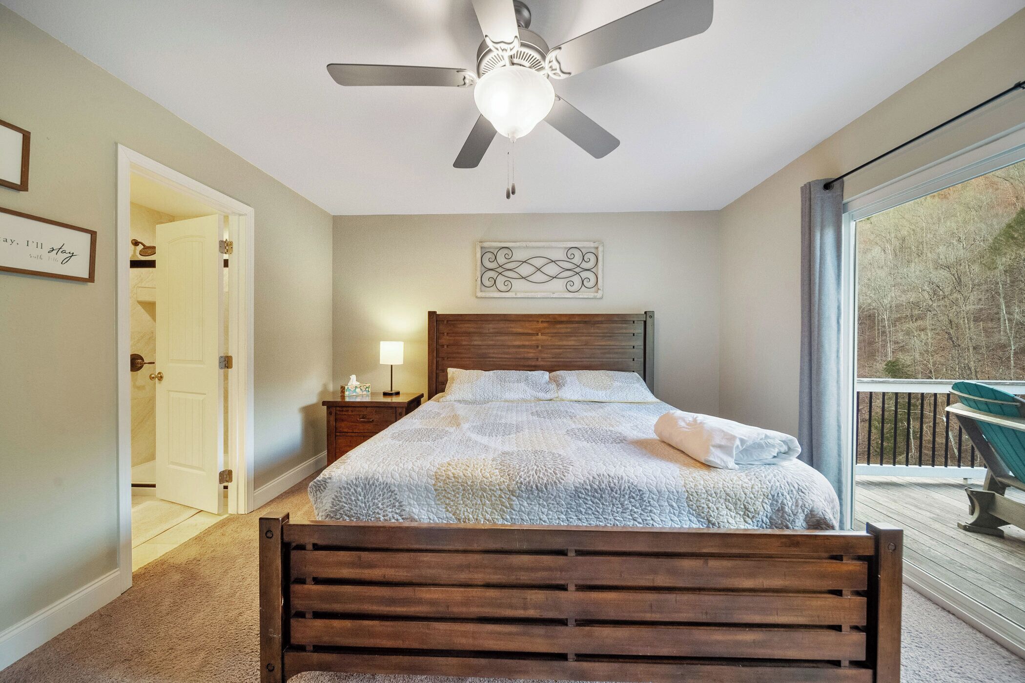 Un dormitorio con cama, mesita de noche, ventilador de techo, ventana con vistas y puerta que da a otra habitación.