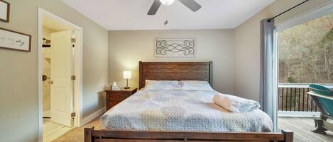 Un dormitorio con cama, mesita de noche, ventilador de techo, ventana con vistas y puerta que da a otra habitación.