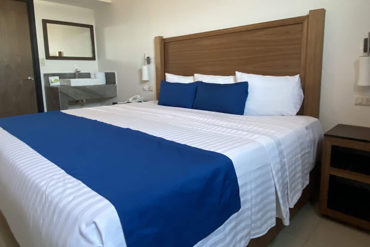 Ein Hotelzimmer mit einem großen Bett, einem Holzkopfende, einem Nachttisch, einem Spiegel und einem Badezimmer mit Waschbecken und Handtuchhalter.