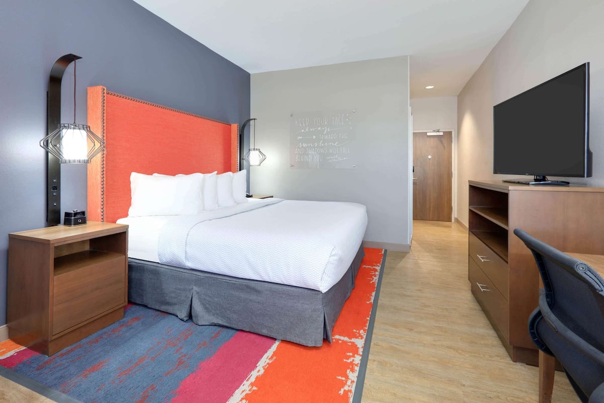 A modern hotel room with a large bed, a desk with a flat-screen TV, a bedside lamp, and a motivational quote on the wall.