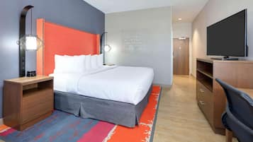 A modern hotel room with a large bed, a desk with a flat-screen TV, a bedside lamp, and a motivational quote on the wall.