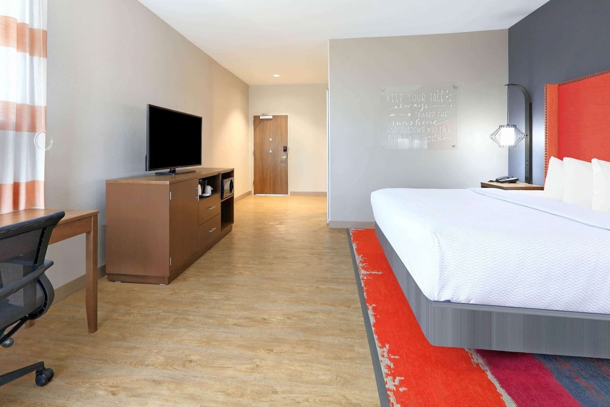 A hotel room with a bed, a desk with a TV, a wardrobe, and a motivational quote on the wall.