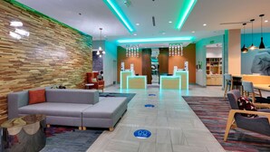 A modern hotel lobby with a wooden wall, a grey sofa, a reception desk, and a dining area. - La Quinta Inn & Suites by Wyndham Lakeway (Lakeway)