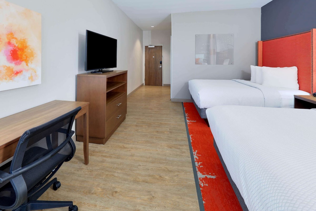 A hotel room with a wooden desk, a TV, two beds, and a red accent rug.