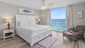 A bedroom with a large bed, a ceiling fan, a view of the ocean, and a wicker chair.