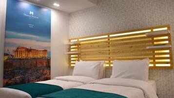 Une chambre d’hôtel avec une grande affiche de l’Acropole d’Athènes, une tête de lit en bois dotée d’un éclairage intégré et deux lits avec du linge de lit blanc et des coussins décoratifs verts.