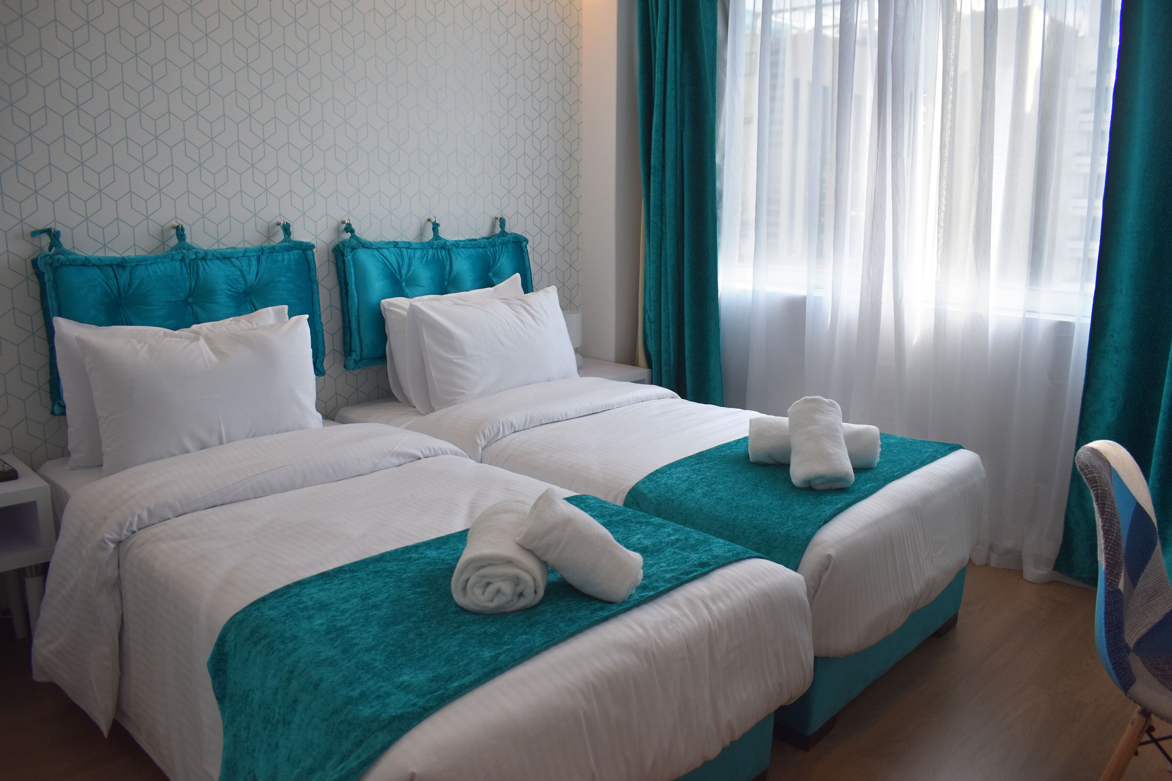 A hotel room with two beds, each with a teal bedspread and white bedding. There are two white pillows on each bed, and a white towel is placed on the bedspread. The room has a window with sheer white curtains and a teal curtain on the side.