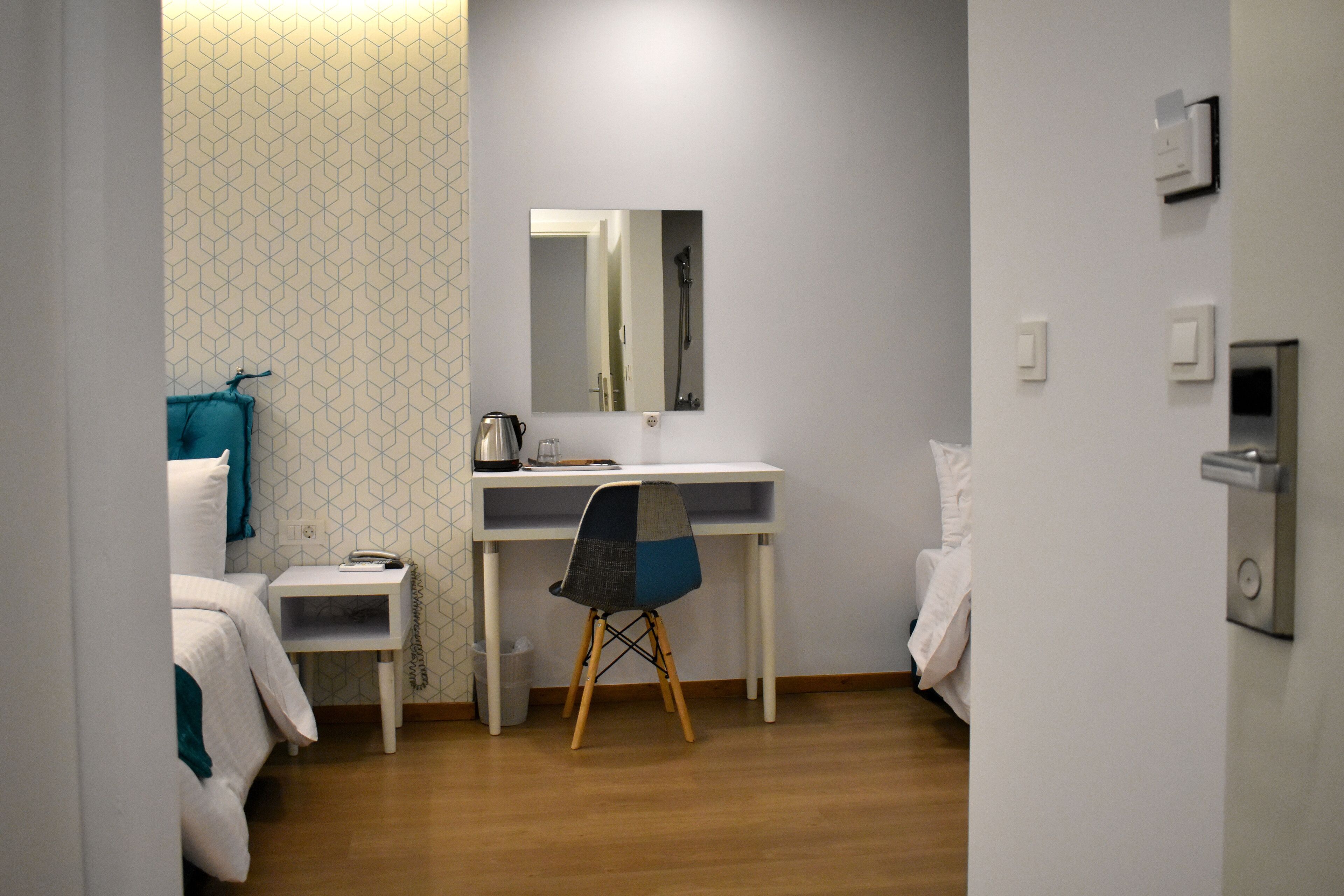 A small, modern room with a wooden floor, a white desk, a chair, a bed, a mirror, and a wall with a geometric pattern.