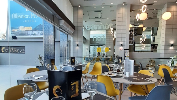 A modern restaurant interior with tables set for dining, a menu stand, and a view of the outside. - Athenian Montaza Hotel (Kallithea)